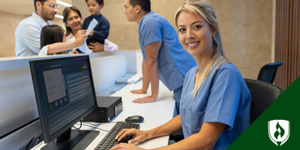 A healthcare worker sits at a front desk in blue scrubs